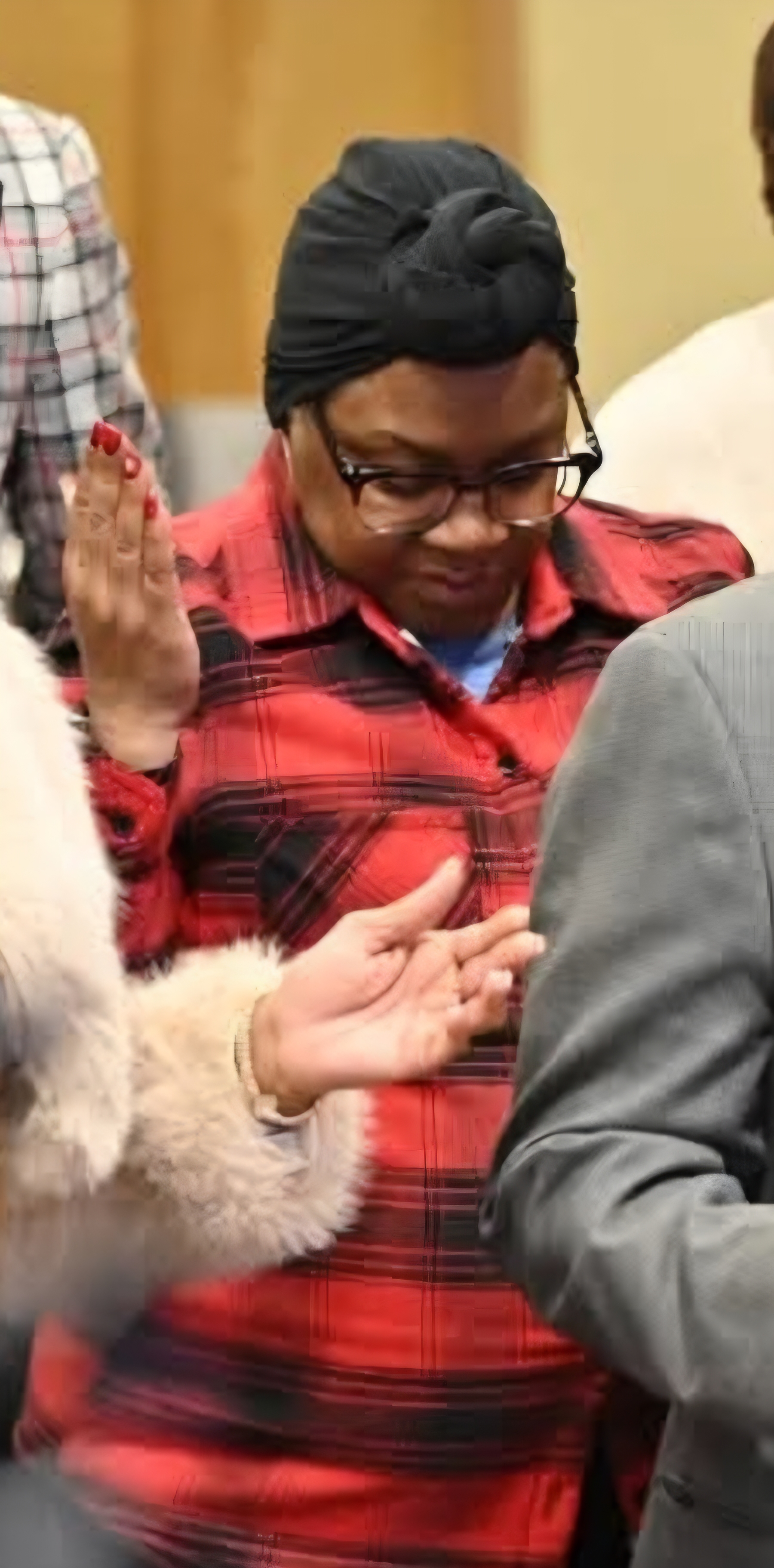 A woman in a red plaid shirt and head wrap is engaged in prayer, with her hands raised, surrounded by others in a church setting.
